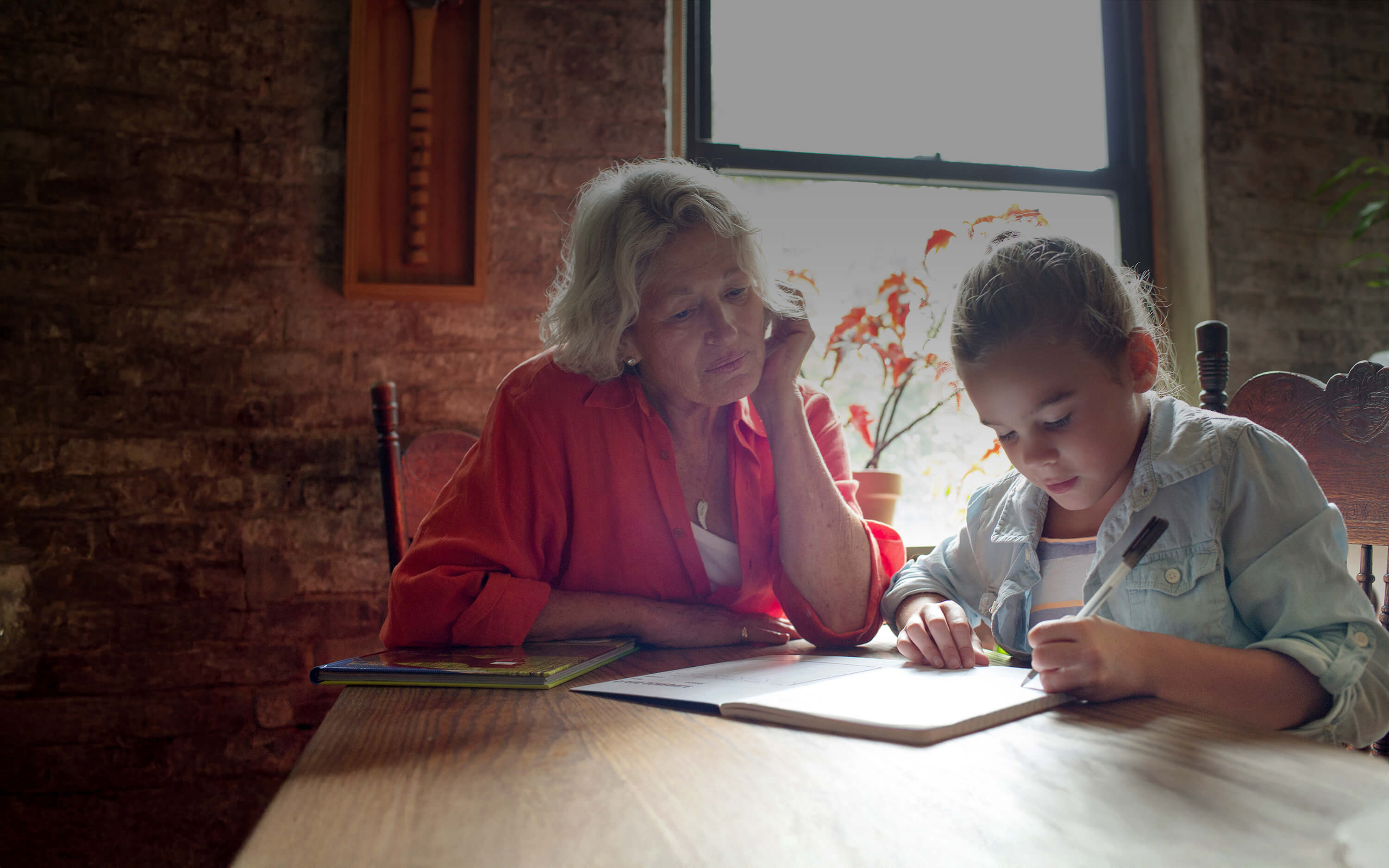 An older woman sitting at a table with a young girl while she's doing homework