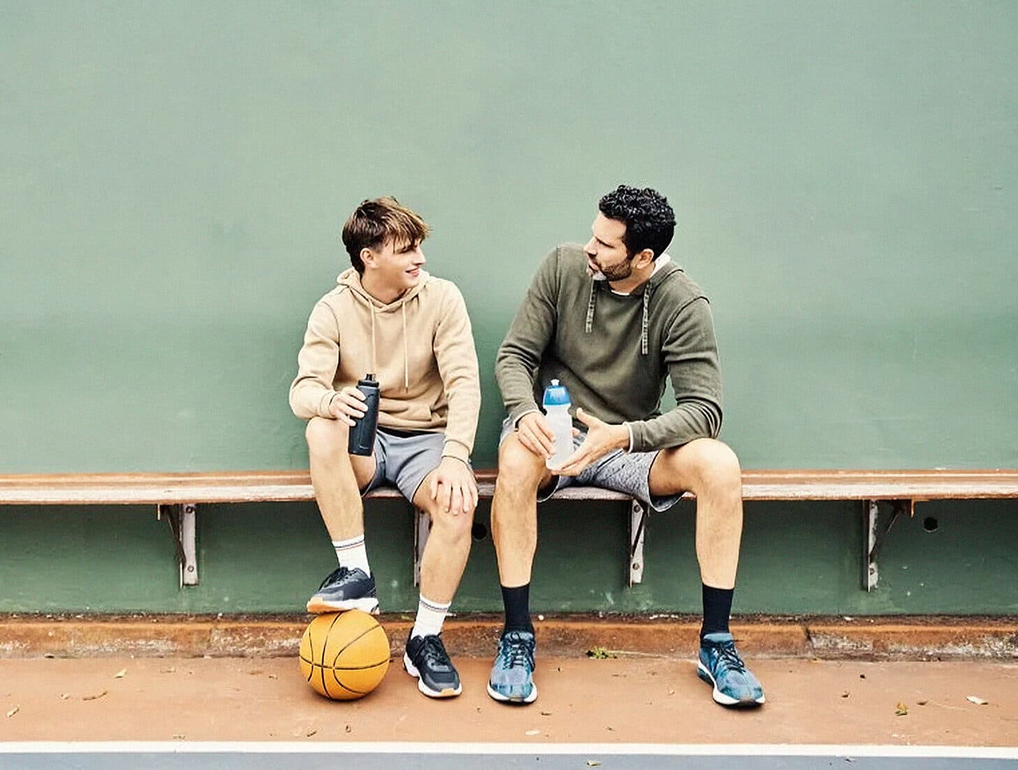 Teenage boy with basketball under his foot looking at father while sitting on a bench seat together